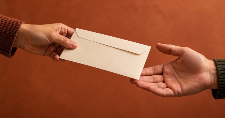 Twee handen die een crèmekleurige envelop overhandigen tegen een terracotta achtergrond.